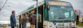 Three individuals stand near a bus marked "Durham Region Transit," waiting at a bus stop on a cloudy day.