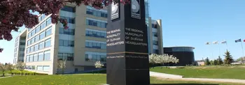 A large sign for "The Regional Municipality of Durham Headquarters" stands on a grassy area in front of a modern office building. The sign displays the address 605 Rossland Road East. Surrounding the area are blooming trees and several flags.