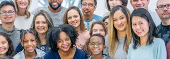 A group of diverse, smiling individuals stands closely together, looking at the camera in a brightly lit space.