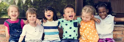Six young children sitting side by side outdoors with arms around each other, wearing colourful clothing and smiling, with a building and greenery in the background.