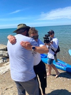 Mike Shoreman hug on shore man Mike Shoreman receives a hug from a man on shore with a camera behind them