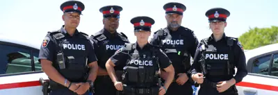 A group of five Durham Region police officers in uniform stand in front of police cars. They are outdoors on a clear day. The officers are wearing caps and their uniforms display the word "POLICE.