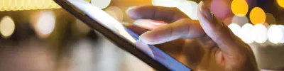 Close-up of hands interacting with a smartphone screen in a brightly lit, blurred urban environment.