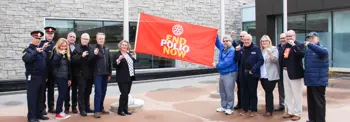 A group of people standing outside a building holds a red banner that reads "End Polio Now." The individuals are gathered in a semi-circle, some making a hand gesture. The background features a modern stone building with large windows.
